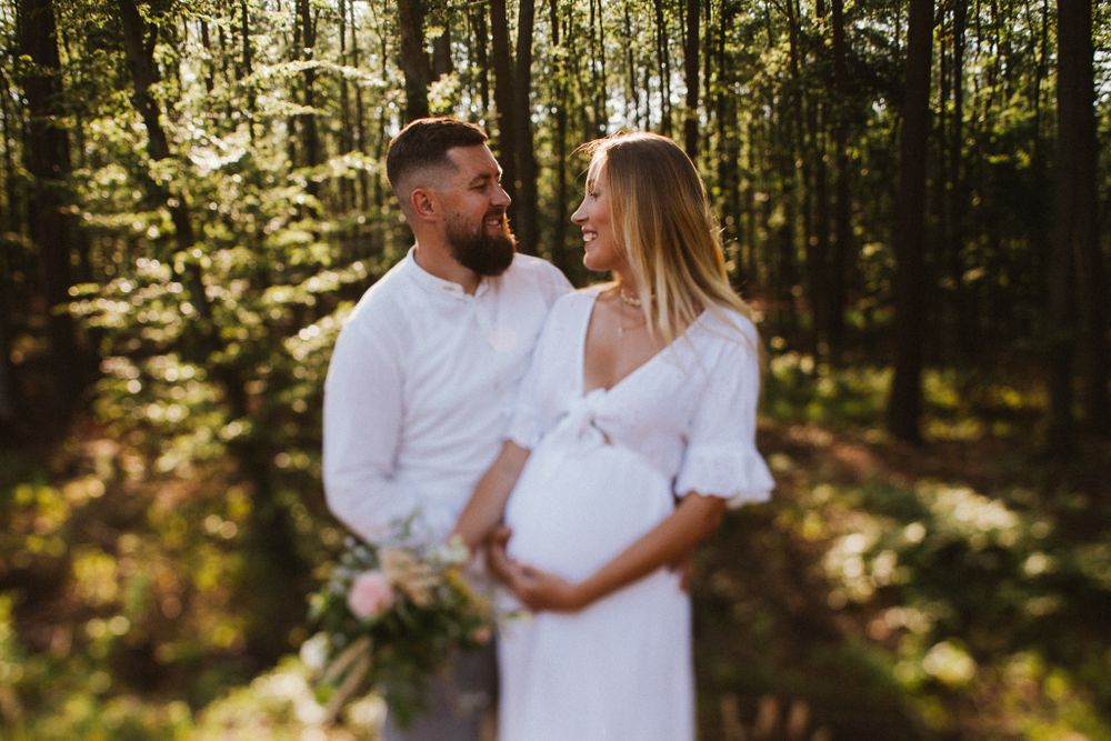 Smiling couple in forest, pregnant woman in white dress holding bouquet.