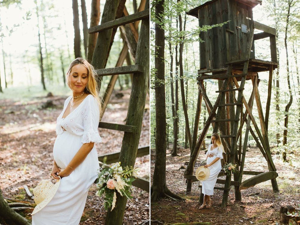 Pregnant woman in white dress standing near wooden hunting tower in forest.