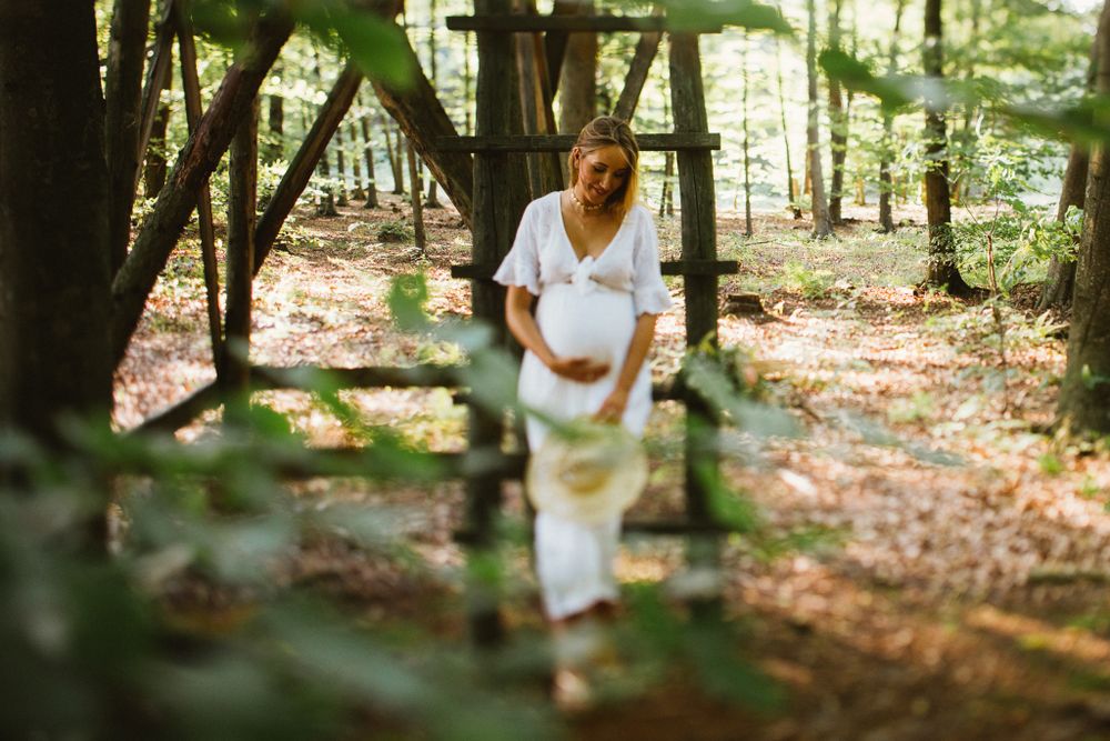 Pregnant woman in white dress cradling belly in sunlit forest.