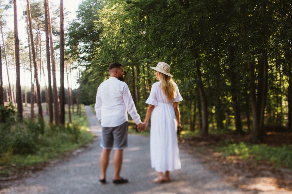 Couple holding hands walking on forest path in white outfits.