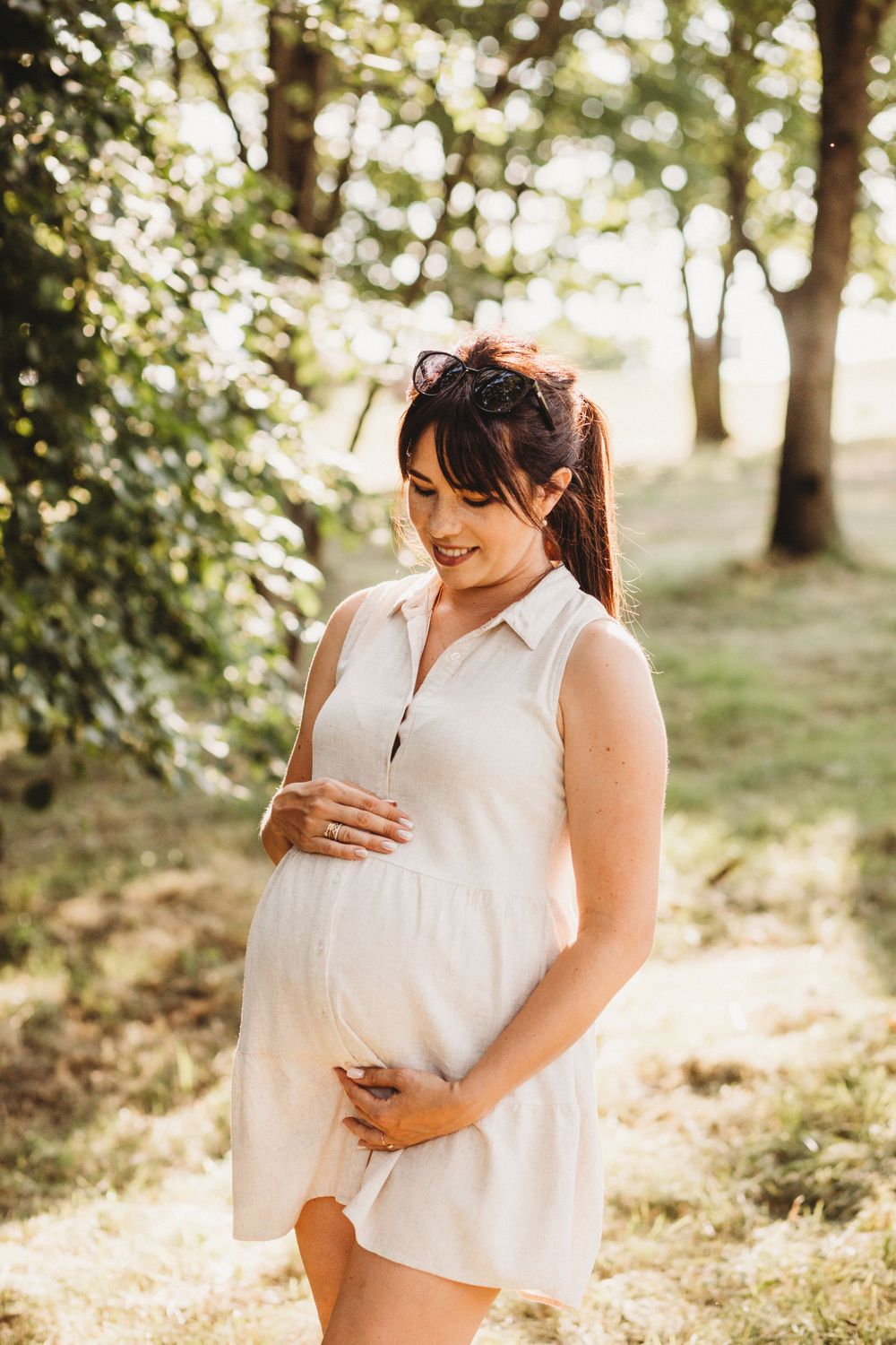 Smiling pregnant woman in white sleeveless dress holding baby bump while standing in sun-dappled park.