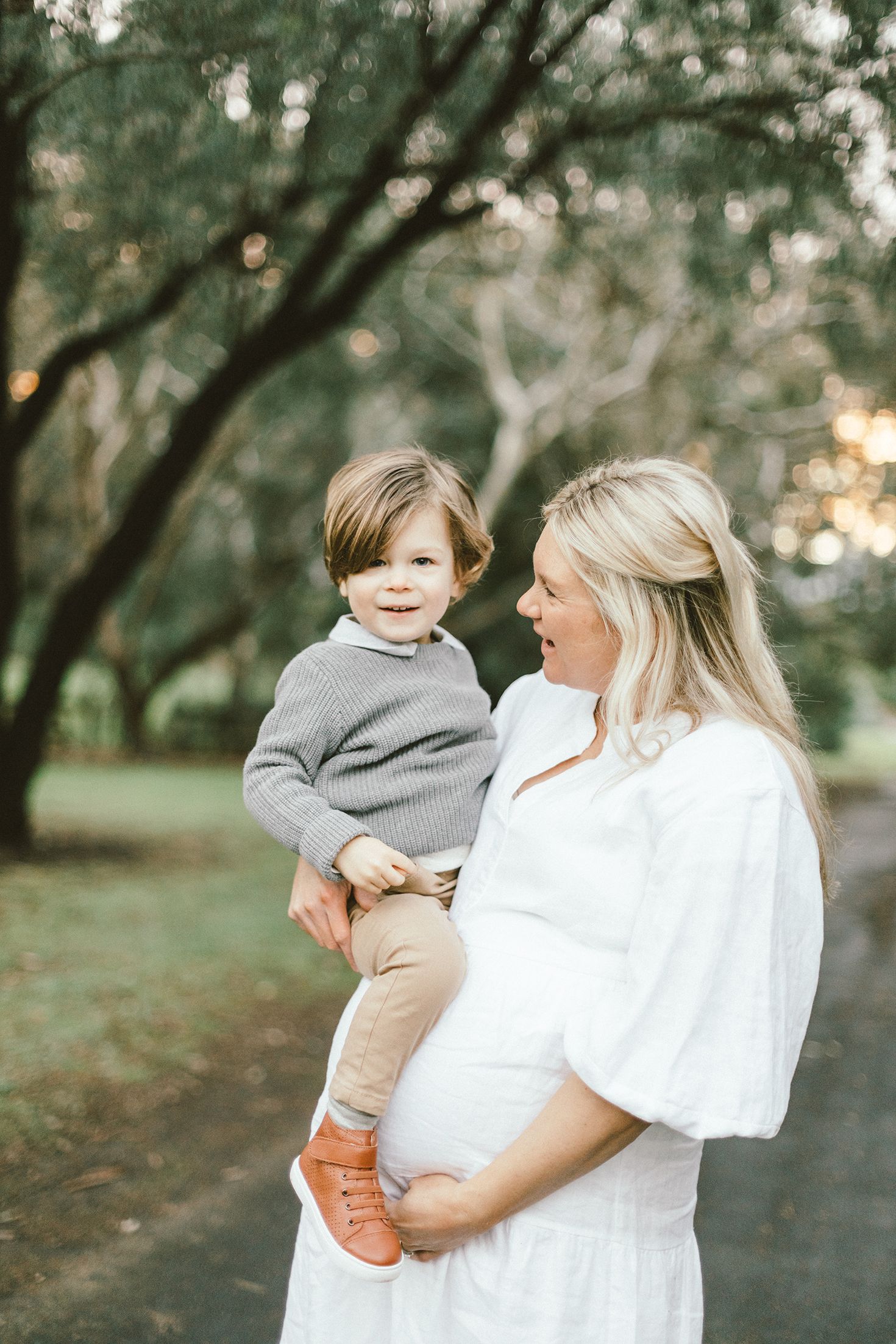 An expectant mother in white holds her child against a backdrop of trees at sunset.