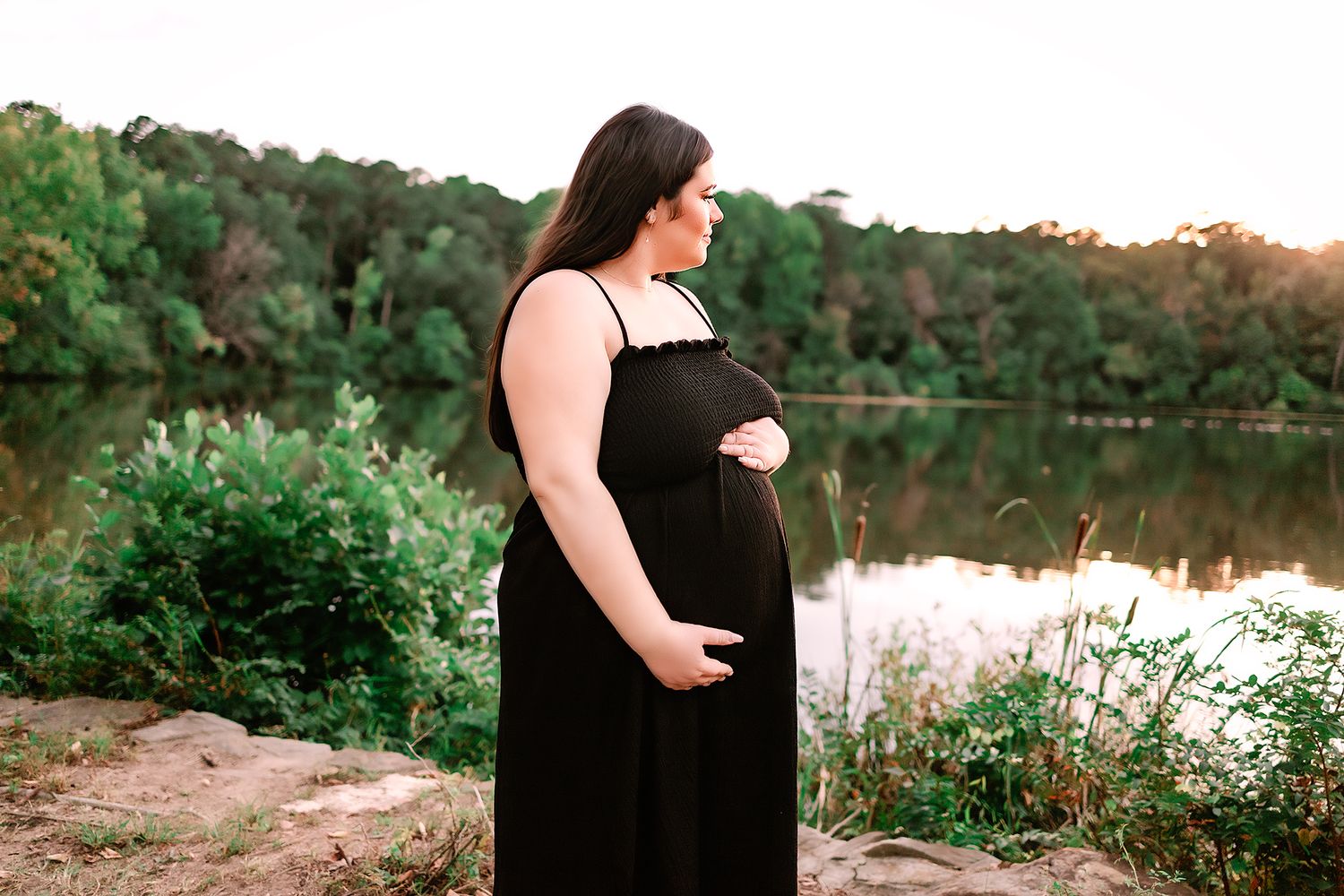 Pregnant woman in a black dress cradling her baby bump near a serene lake at golden hour.