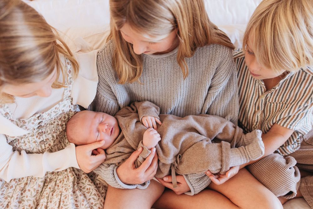 Three children gathered around a newborn baby wearing a gray onesie on white bedding.