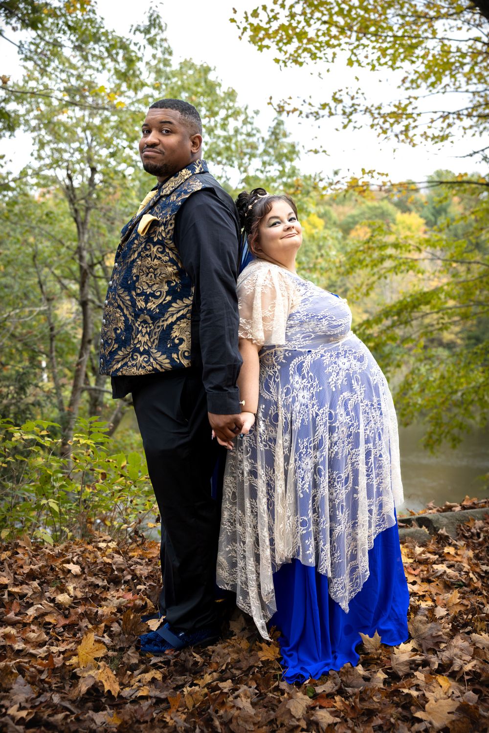Couple stands back-to-back in elegant attire, holding hands in a forest setting with autumn leaves and a lake in the background.