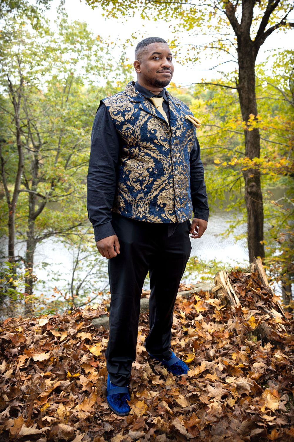 A groom wearing an ornate gold and navy vest stands in autumn foliage near a river.