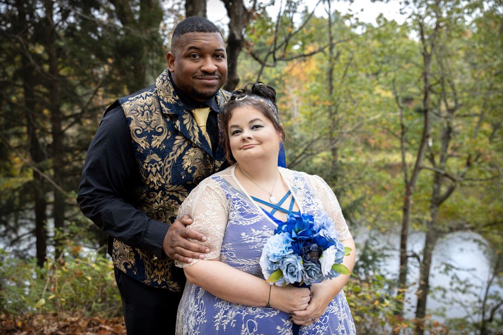 A wedding couple poses together in formal attire against a scenic autumn backdrop with a river.