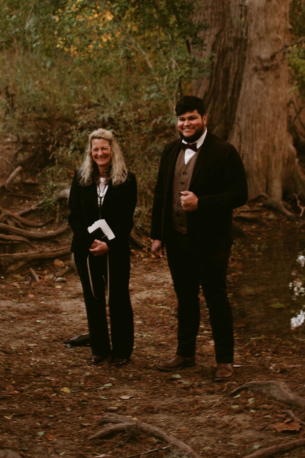 A couple stands together on a forest path surrounded by autumn trees.