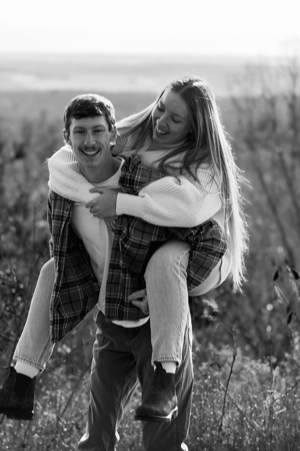 A playful couple shares a joyful moment in a black and white outdoor portrait.