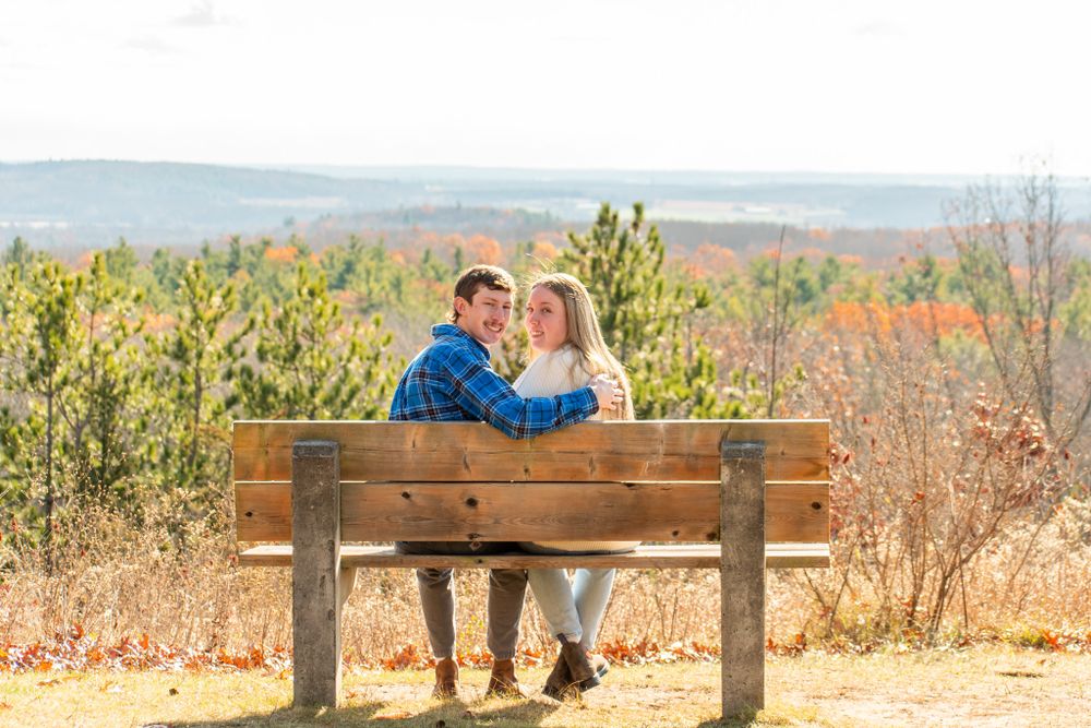A couple sits close together on a wooden bench overlooking rolling hills in autumn.