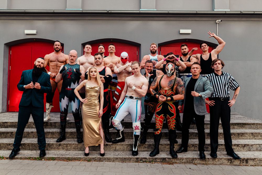 A group of wrestlers and performers pose together on steps in front of red doors for a group photo.