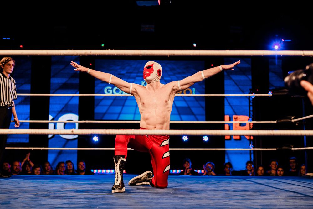 A wrestler in red pants strikes a dramatic pose with arms outstretched in a wrestling ring under blue lighting.