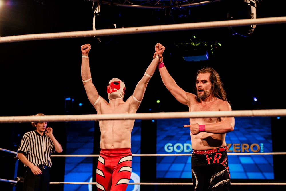 Two wrestlers embrace in victory after a match in a brightly lit wrestling arena.