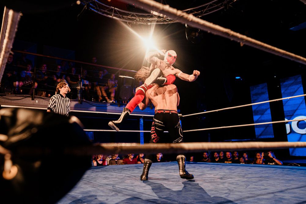 A wrestling match in progress shows intense action between competitors in a brightly lit ring.