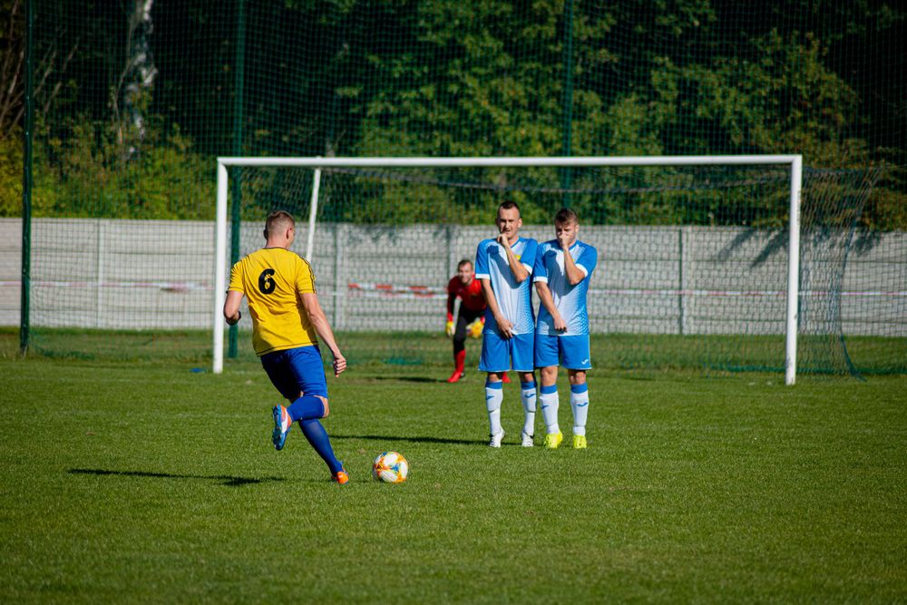 Soccer players compete on a sunny green field with goal posts visible in the background.