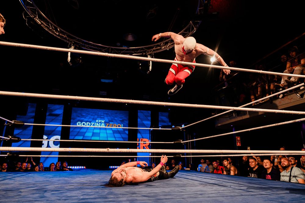 A dramatic aerial wrestling move being executed above a fallen opponent in the ring.