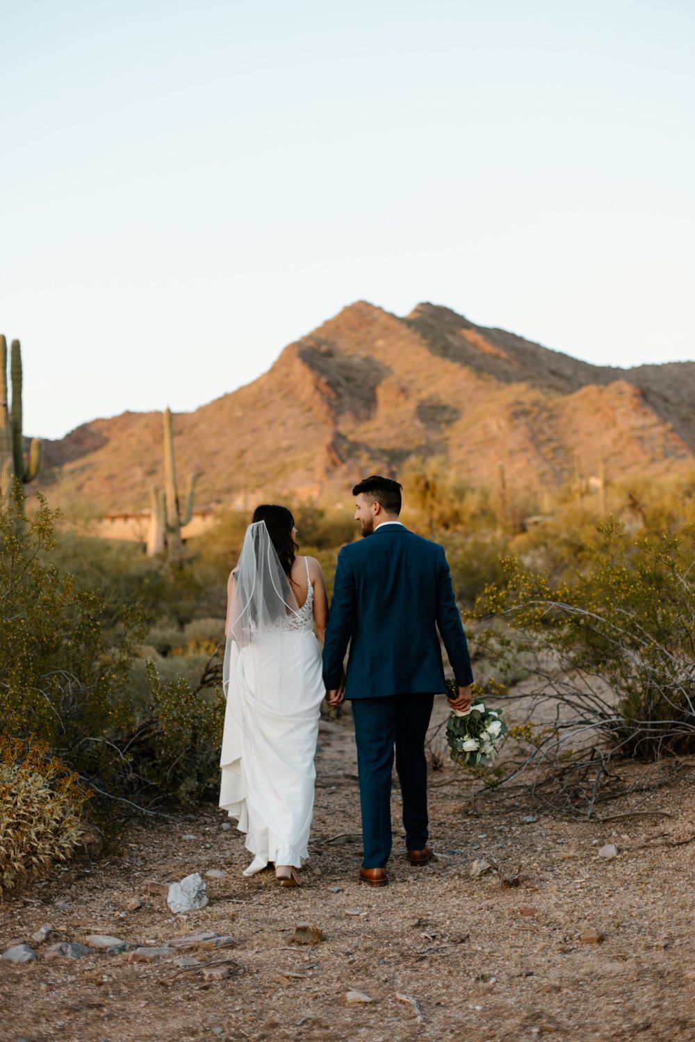Bride and groom walk along desert path with bouquet against mountain landscape and saguaro cactus.