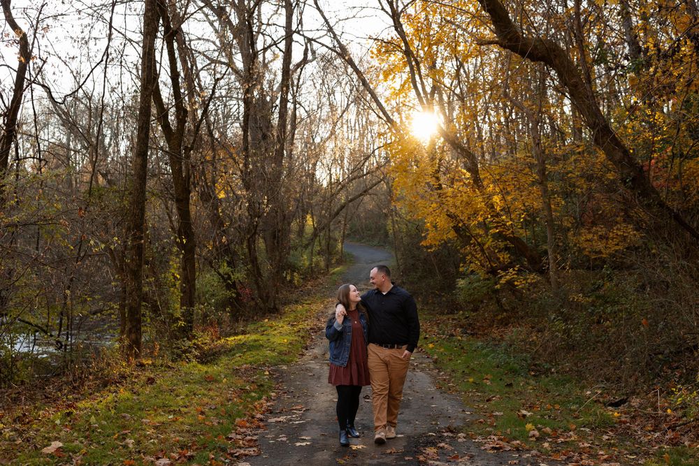 A couple walks hand in hand on a trail during golden hour with autumn trees surrounding them.