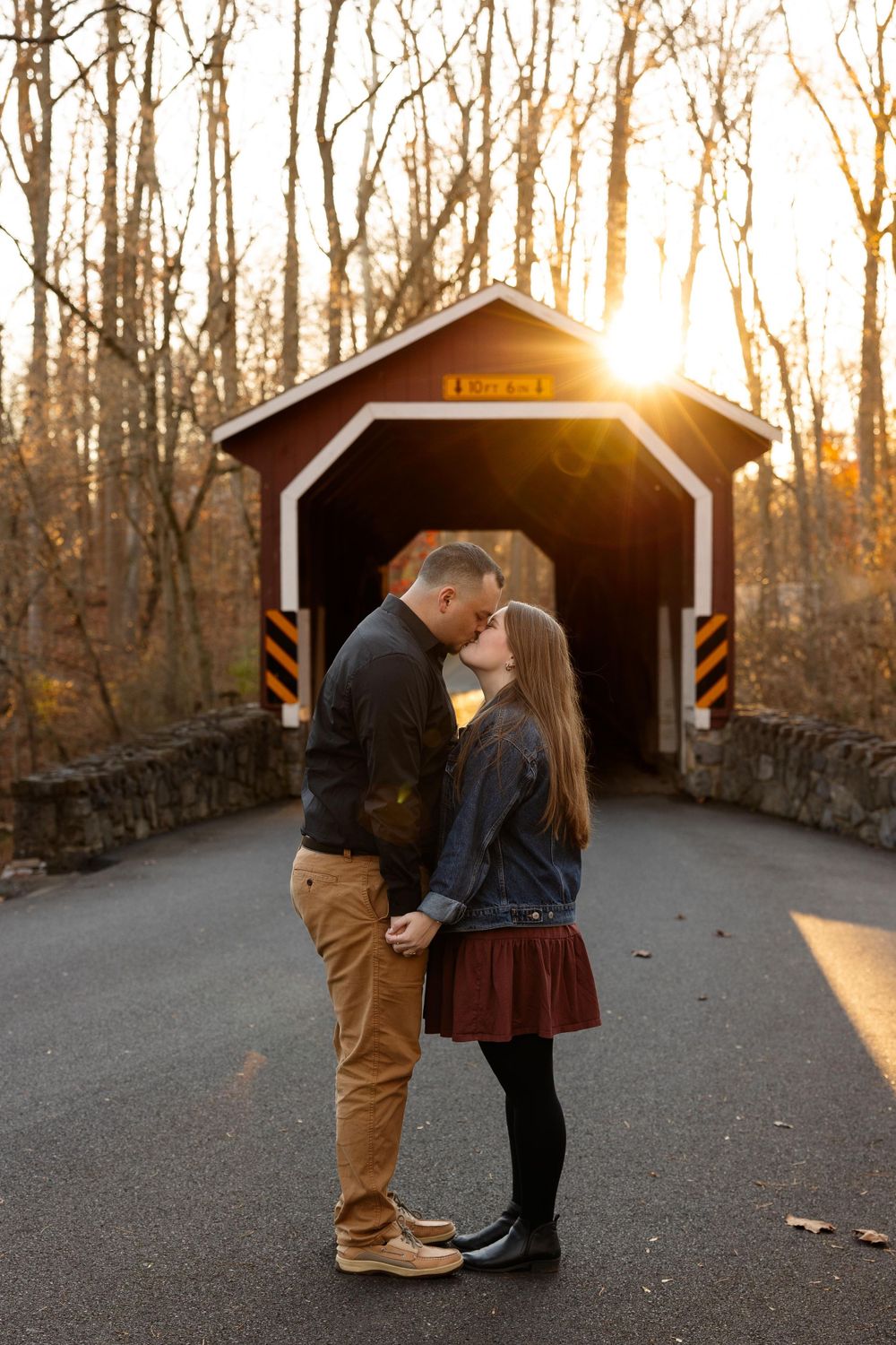 A couple shares a romantic moment in front of a covered bridge during golden hour sunset.