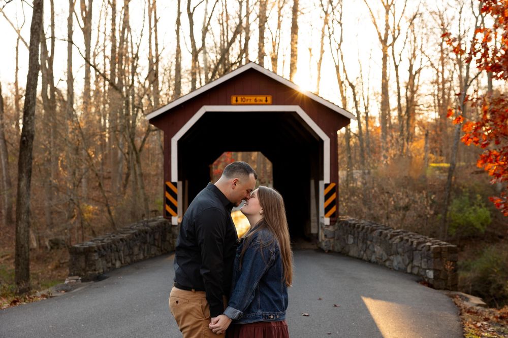 A couple embraces in front of a rustic covered bridge surrounded by autumn trees.