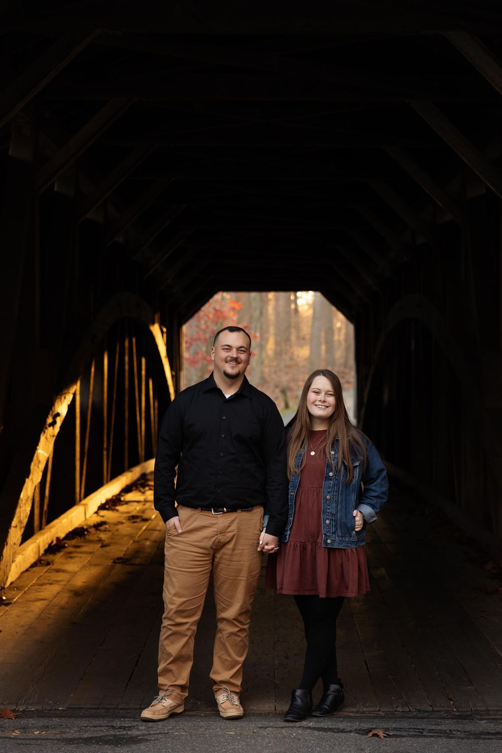 A couple stands holding hands inside a covered bridge with autumn foliage visible in background.