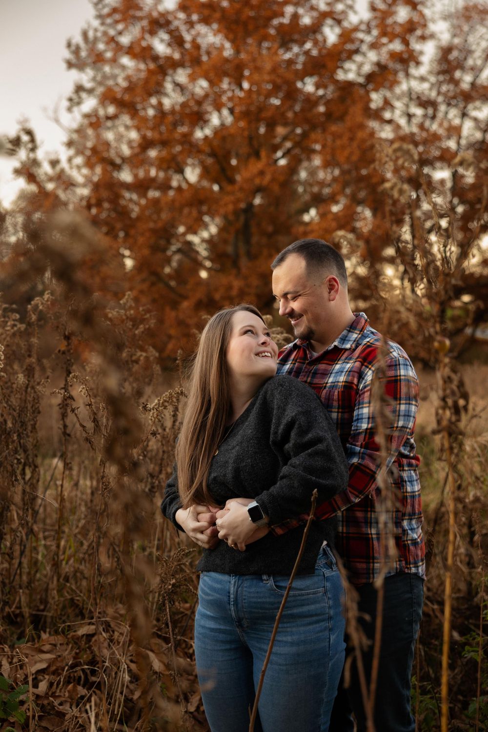 A romantic autumn engagement photo showing a couple embracing in a field with orange trees in the background.