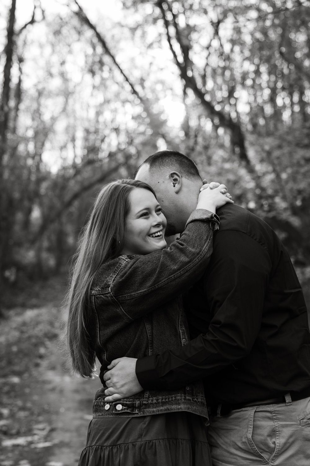 A romantic black and white portrait of a couple embracing and laughing in a wooded setting.