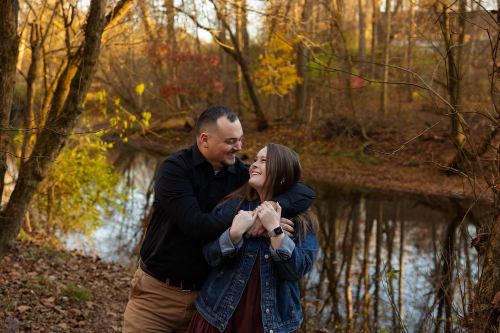 A couple shares an intimate moment by a pond during autumn with golden sunlight filtering through trees.