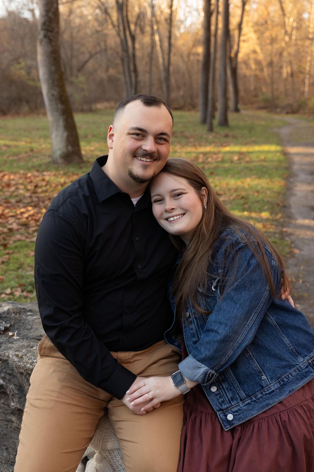 A couple in casual attire poses together on an autumn day in a park setting.
