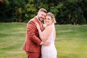 Joyful outdoor wedding portrait with rust-colored suit and lace white dress.