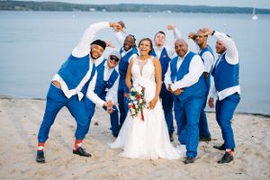 Wedding party poses playfully on a beach in coordinated blue and white attire by the waterfront.