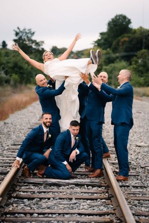 Wedding party creates playful lifting pose on railroad tracks with navy suits and white dress.