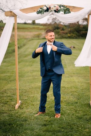Wedding party member adjusts his navy suit beneath a decorated wooden arch on a grassy lawn.