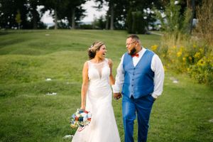 Newlyweds share loving glances while walking together on grassy hillside.