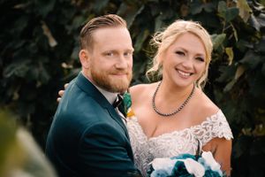 Wedding portrait of couple in navy suit and lace dress with blue bouquet against greenery.