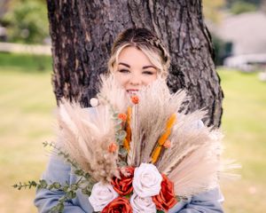 A lovely wedding bouquet featuring red and white roses with pampas grass and dried orange flowers against a tree trunk.