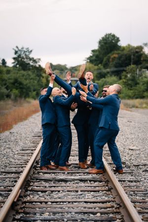 Groomsmen in navy suits playfully lift someone on railroad tracks during wedding photoshoot.