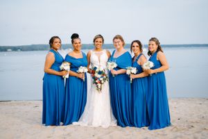 Bridal party in royal blue dresses poses on sandy beach with water view.