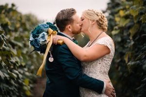 Romantic kiss between newlyweds holding blue bouquet in vineyard row.