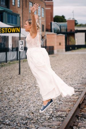 Bride in flowing white dress and sneakers jumps playfully on railroad tracks.