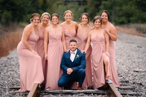 Wedding party in pink dresses and navy suit poses together on railroad tracks.