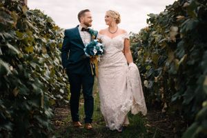 Couple in formal wedding attire walking through vineyard rows with lace dress trailing behind.