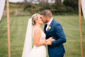 Newlyweds share their first kiss under a wooden arch decorated with white fabric and flowers.