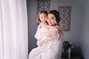 Two people share a tender moment in white lace wedding attire near a window.