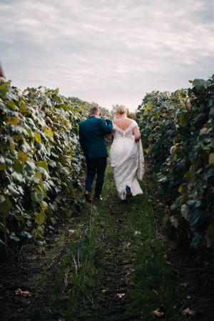 A wedding couple walks through vineyard rows under cloudy skies.