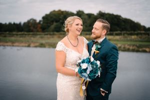 Wedding couple laughs together while holding a teal and white bouquet near a lake.