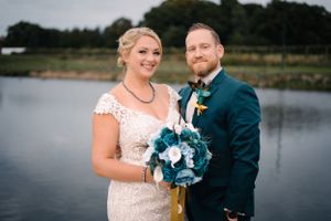 A couple poses by a lake with the bride holding a teal and white bouquet against a navy suit and lace dress.