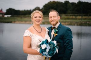 Happy wedding couple poses with teal bouquet against serene lake backdrop.
