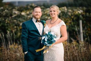 Wedding portrait of couple in formal attire with blue bouquet against natural landscape.