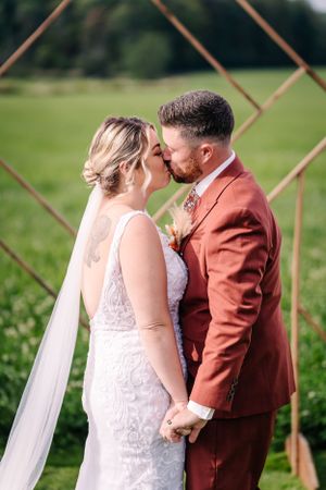 Romantic wedding kiss under a geometric copper arch in a lush green field.